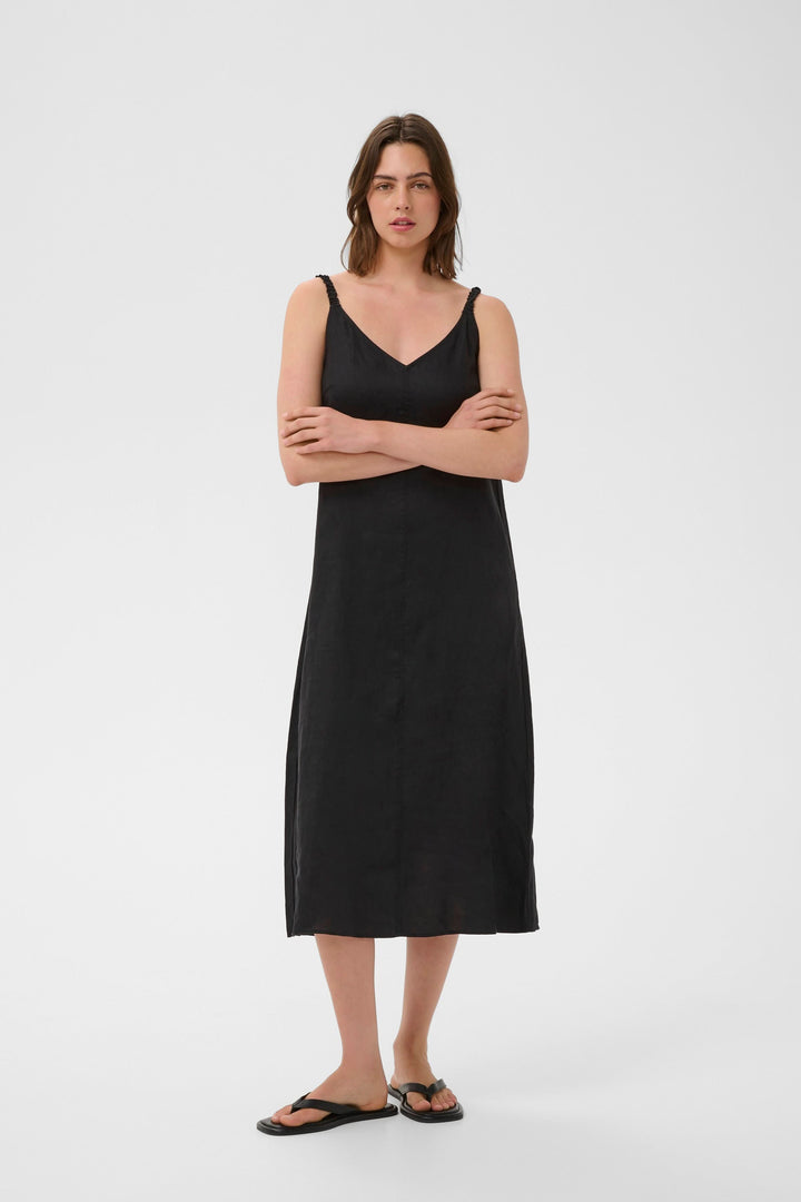 Femme portant la Robe Elinor midi fluide à bretelles fines, en lin noir, avec coupe ample et encolure en V.
