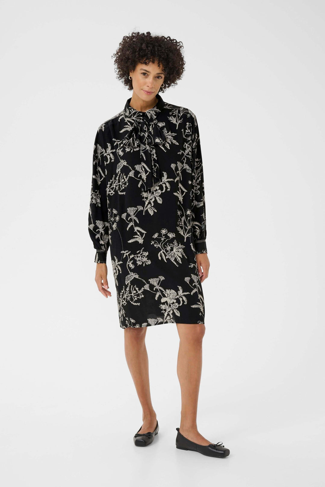 Femme portant une Robe Sanes noire à imprimé floral avec col lavallière et manches longues, style chic et féminin.