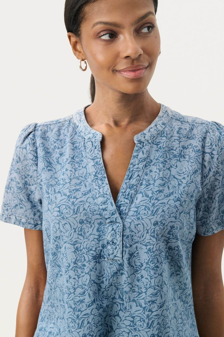 Modèle portant une blouse à manches courtes fleurie en coton bleu, avec un décolleté boutonné.