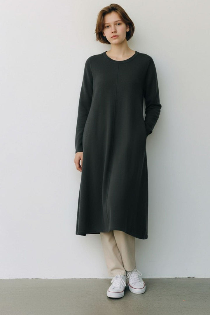 Femme portant une robe de tricot noire à manches longues, avec encolure ronde et ligne verticale, posant debout contre un mur blanc.
