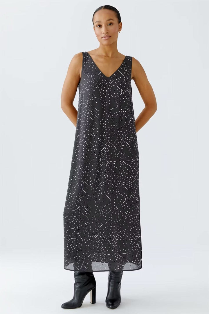 Robe longue sans manches avec imprimé discret à pointillés, encolure en V, et fentes latérales, portée avec des bottes hautes.