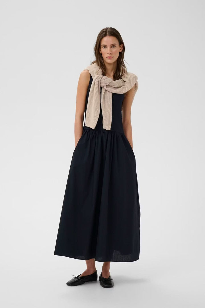 Robe midi sans manches Dagnama, femme portant une robe noire élégante avec jupe ample, style polyvalent et intemporel.