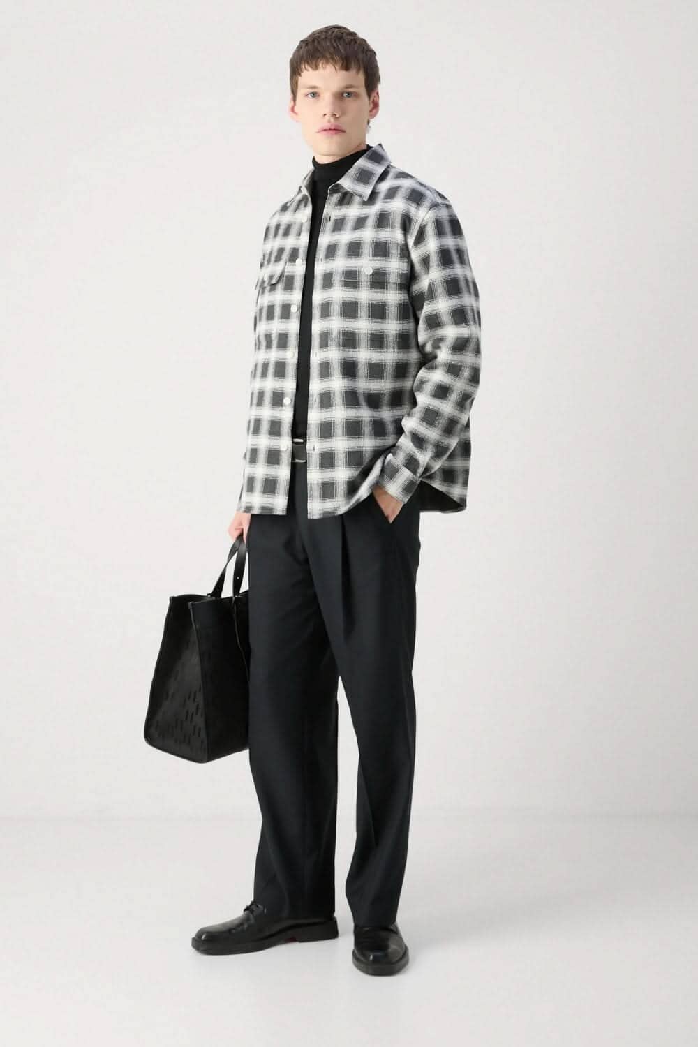 Homme portant une surchemise à carreaux noir et blanc avec un t-shirt noir et un pantalon sobre, tenant un sac noir.