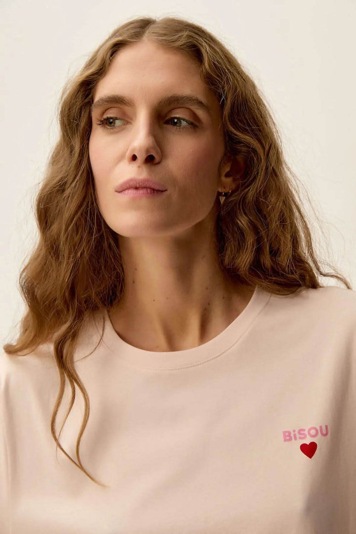 Femme portant un t-shirt rose nude avec imprimé 'Bisou' en cœur, style décontracté et moderne.