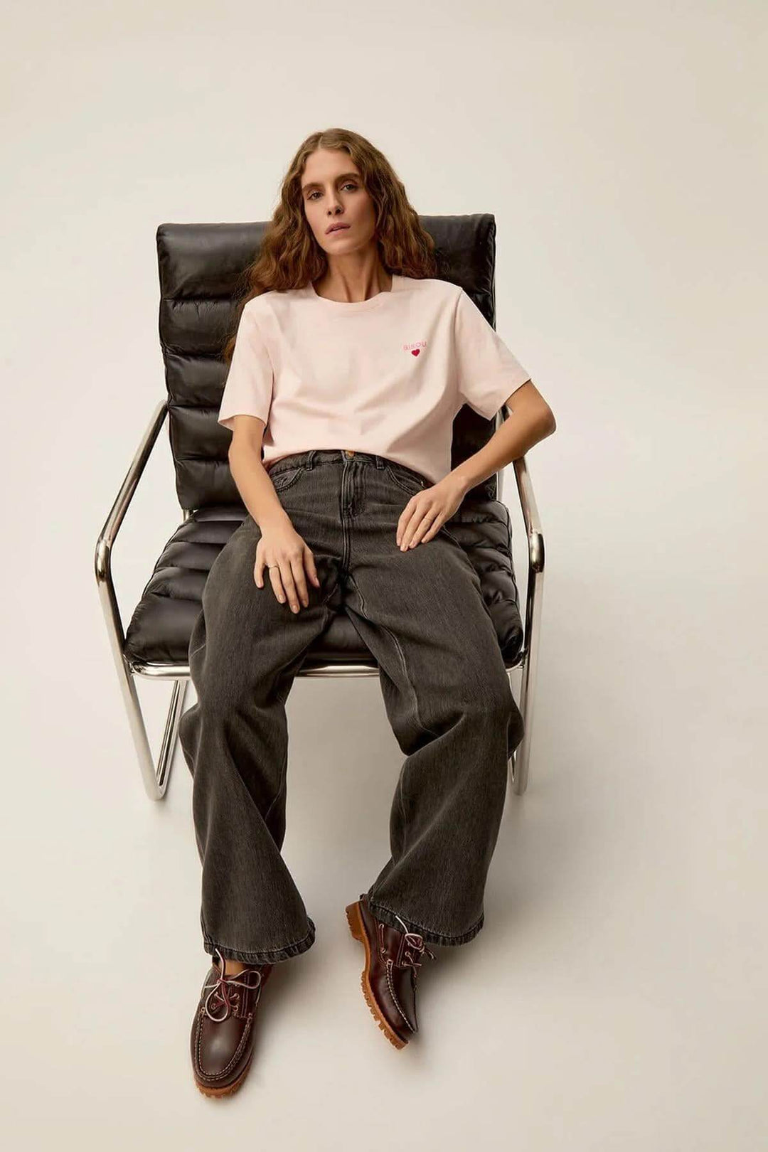 Modèle portant un t-shirt Gazoline rose avec imprimé 'Bisous', assis sur une chaise moderne.