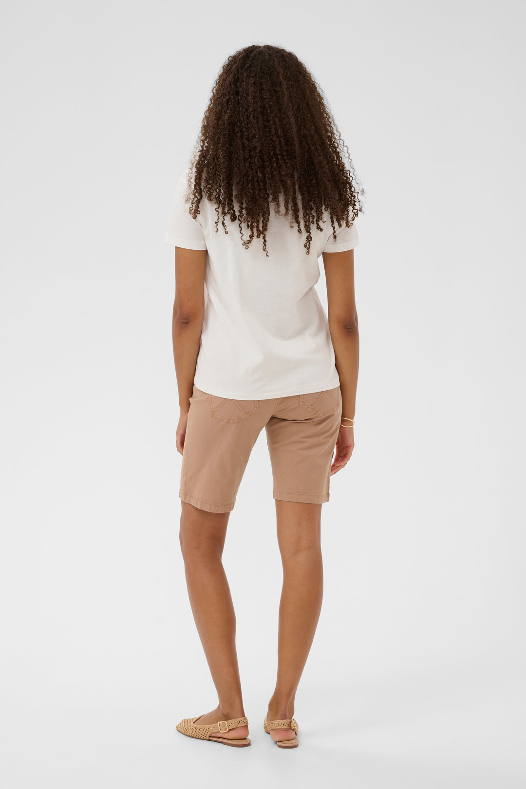 Femme portant un T-shirt Joliy blanc, vue arrière, avec des manches courtes et un short beige, ajoutant une touche décontractée.