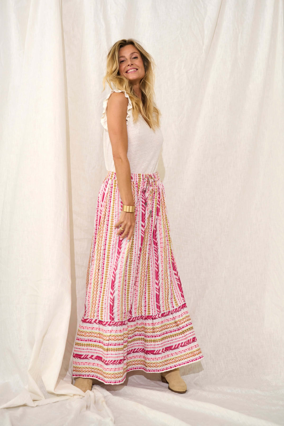 Femme souriante portant un t-shirt blanc sans manches et une jupe longue multicolore à motifs sur fond clair.