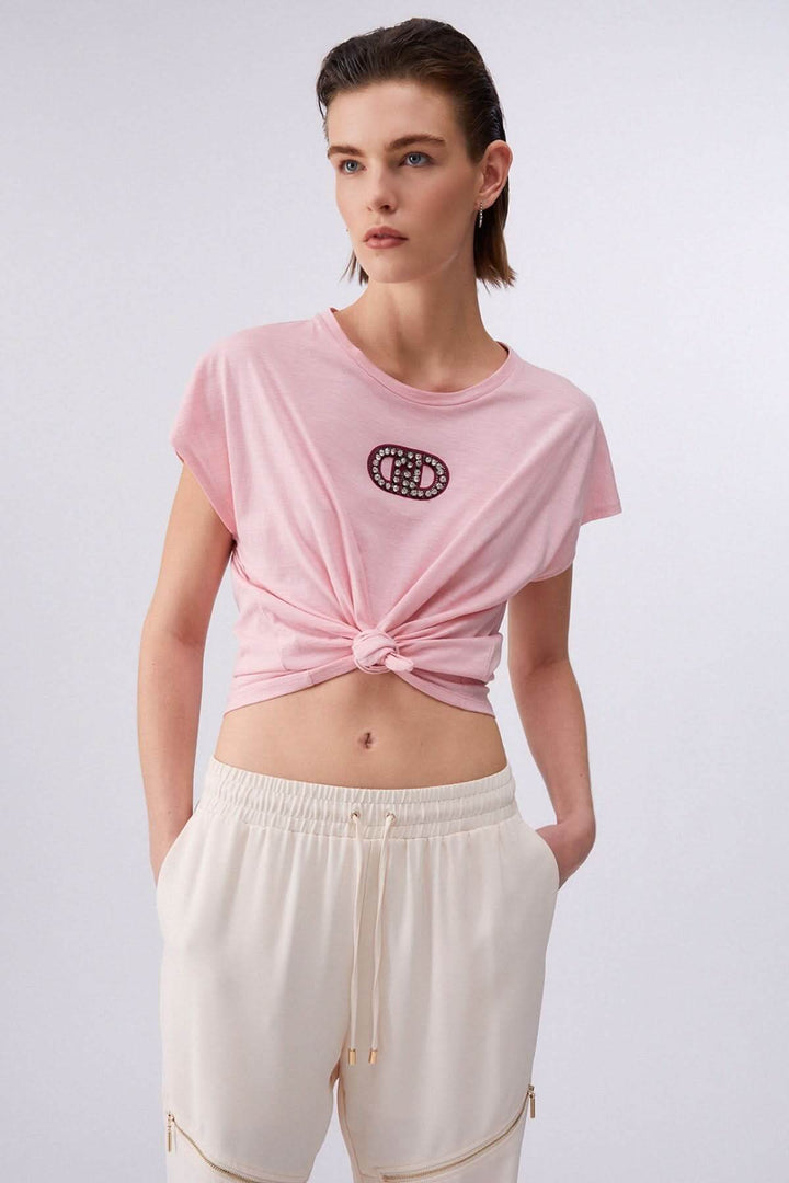 Femme portant un T-shirt avec logo décoratif, coupe décontractée, manches courtes et encolure ronde, associé à un pantalon léger.