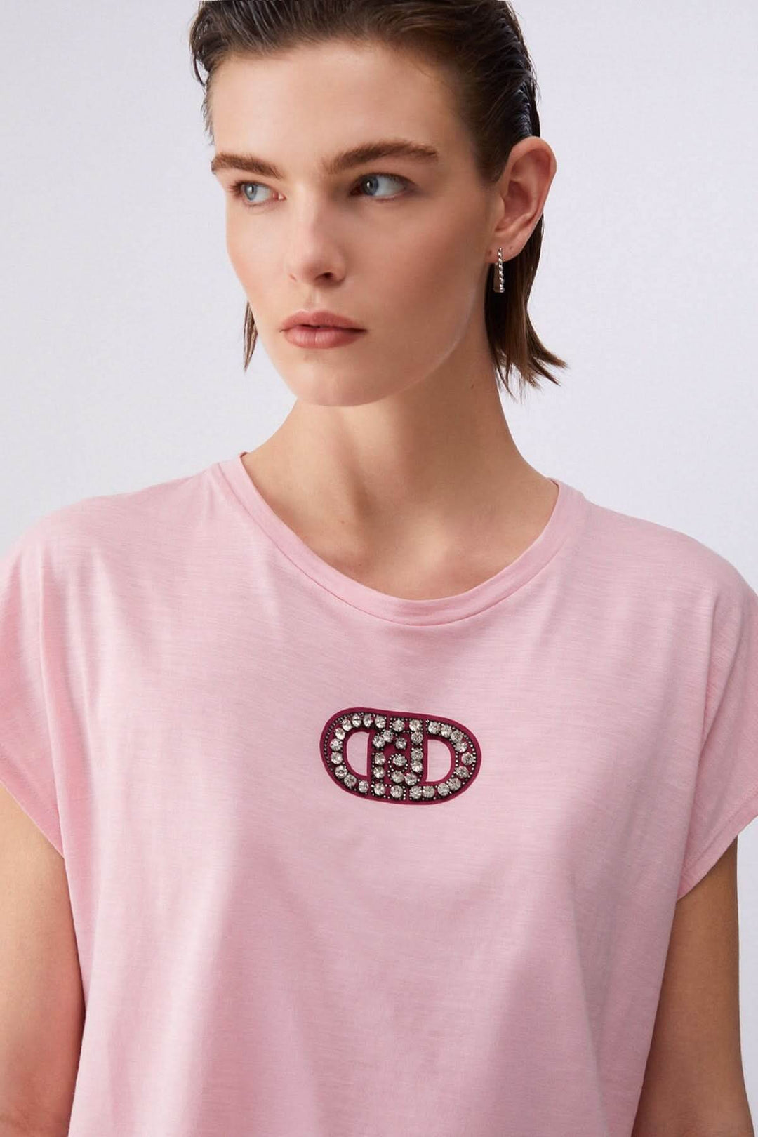 T-shirt avec logo décoratif rose, manches courtes et encolure ronde, porté par un modèle. Tissu doux et confortable pour un style élégant.
