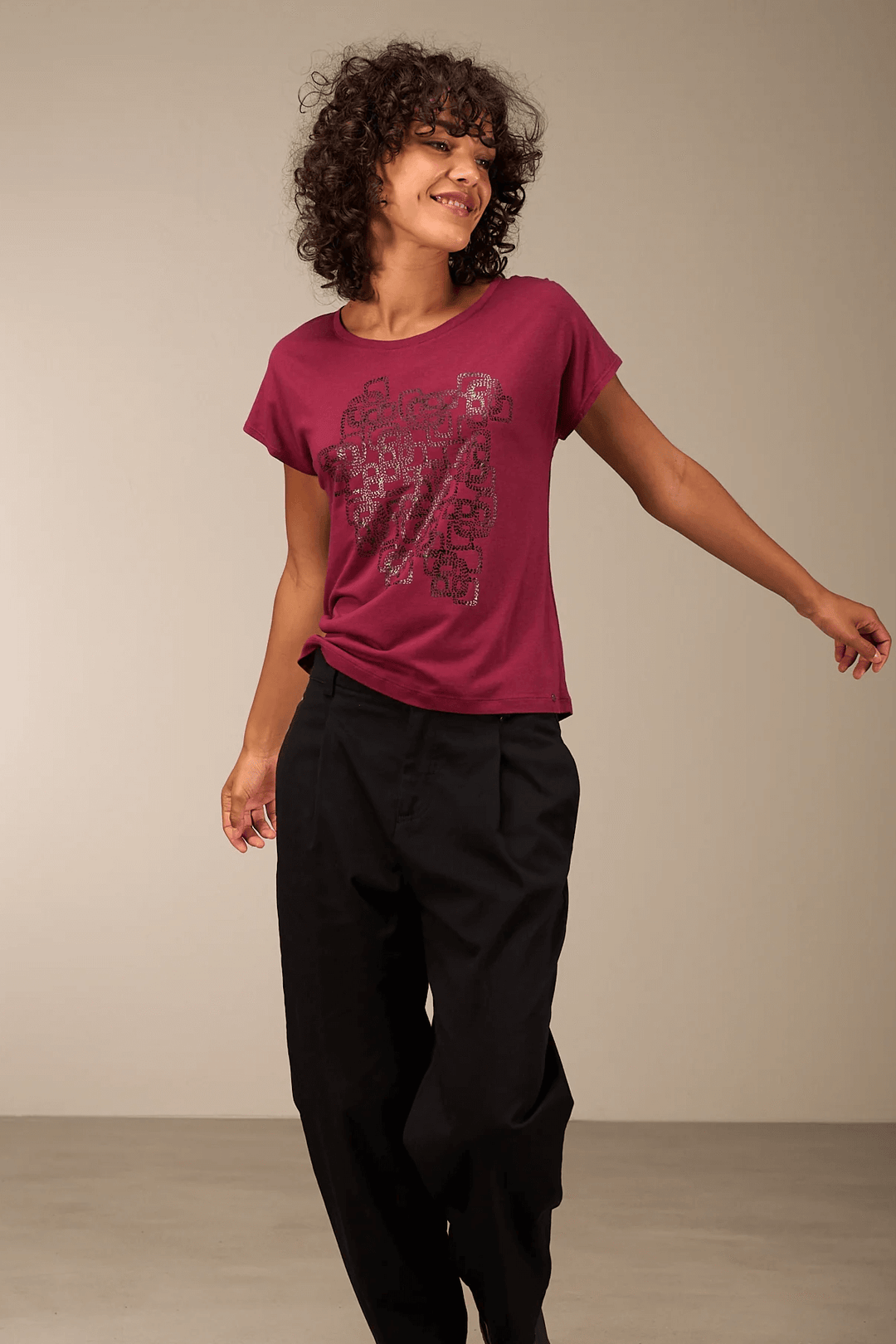 Femme portant un t-shirt imprimé violet avec un motif graphique, manches courtes et col rond, assorti à un pantalon noir, style décontracté.