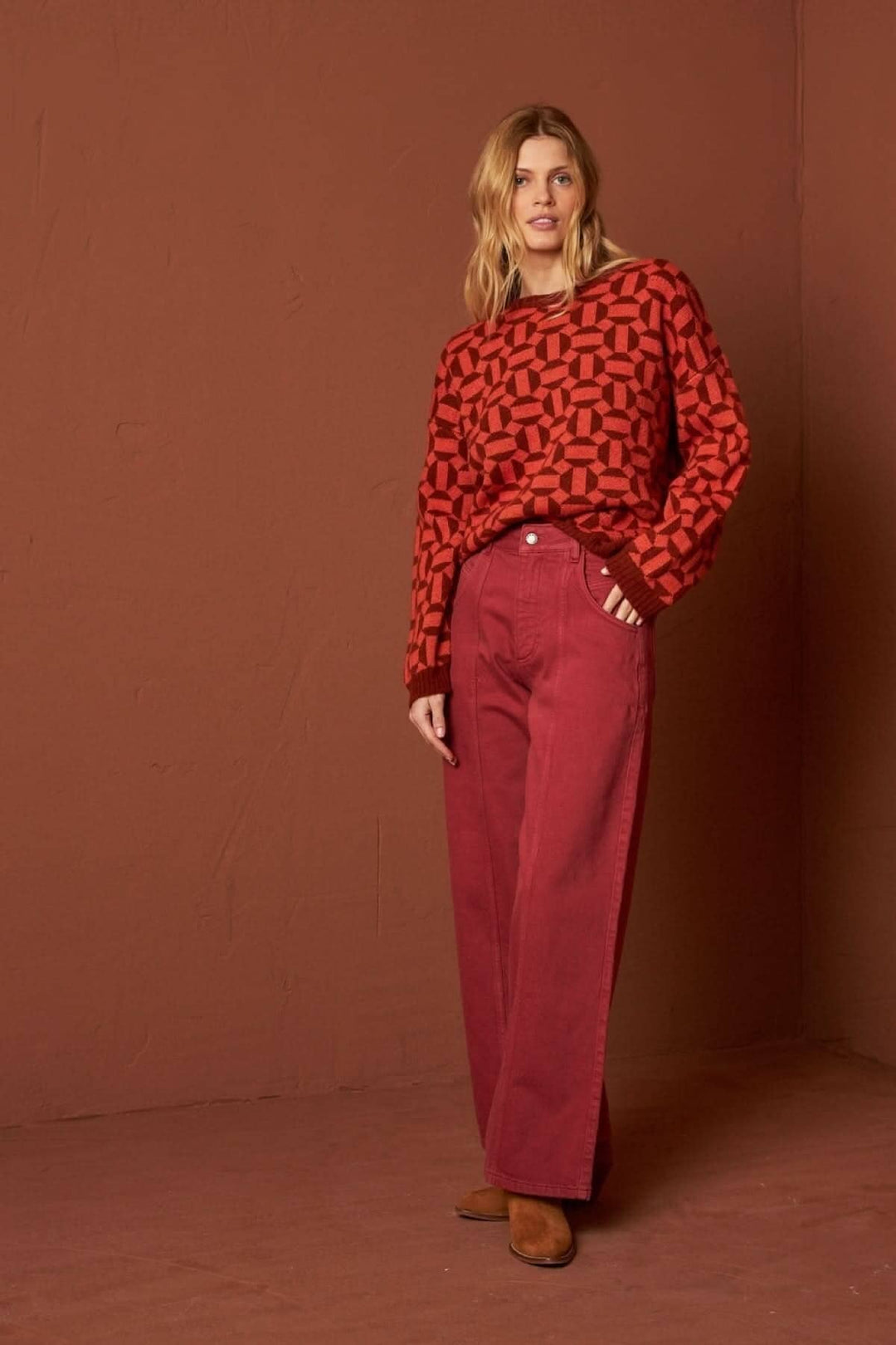 Femme portant un tricot à motifs géométriques rouges et un pantalon large bordeaux, sur fond marron.