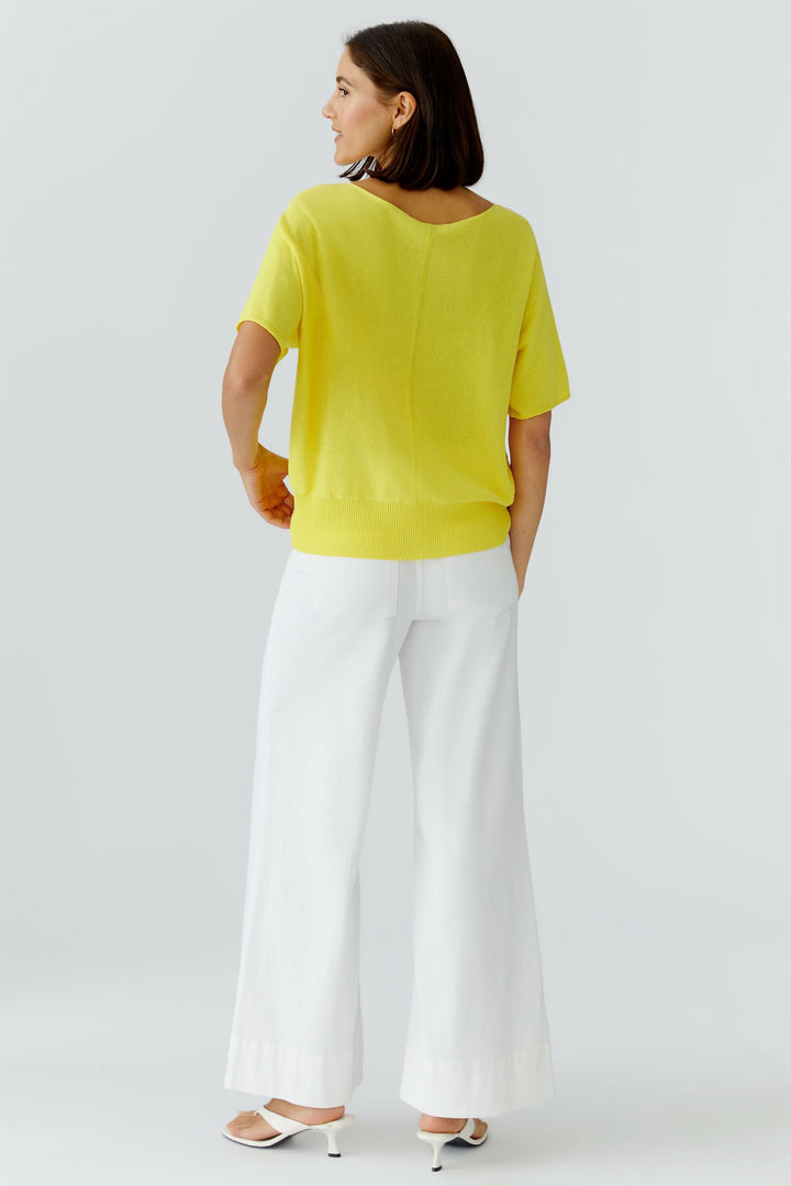 Femme portant un tricot fluide à manches courtes jaune, coupe fluide avec poche poitrine, associé à un pantalon blanc ample et des talons.