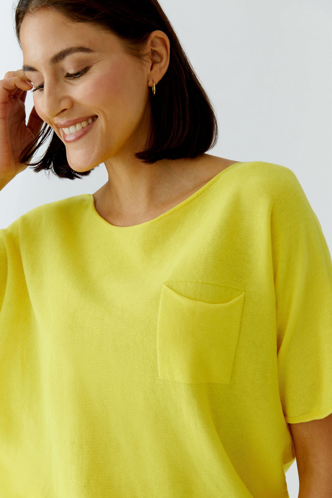 Tricot fluide à manches courtes en jaune, avec encolure dégagée et poche poitrine, porté par un modèle souriant pour un look moderne.