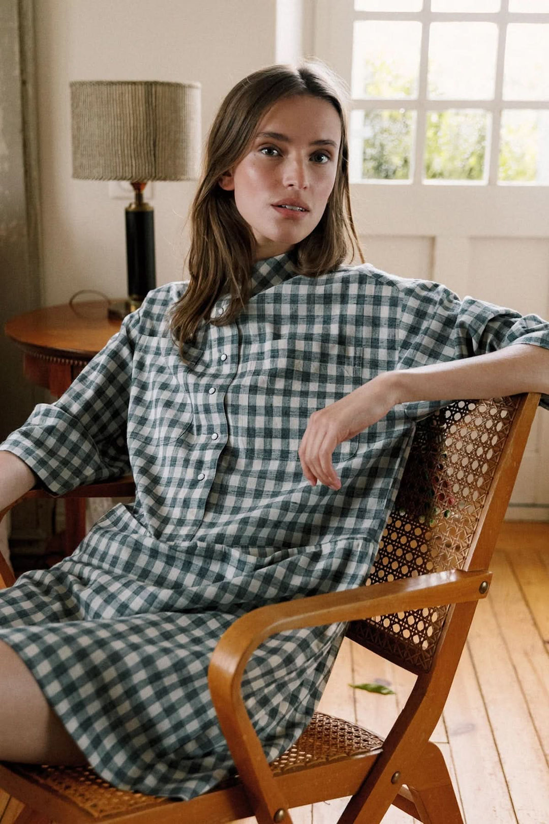 Femme portant une tunique à carreaux Vichy, assise dans une chaise en bois, style décontracté, ambiance intérieure.
