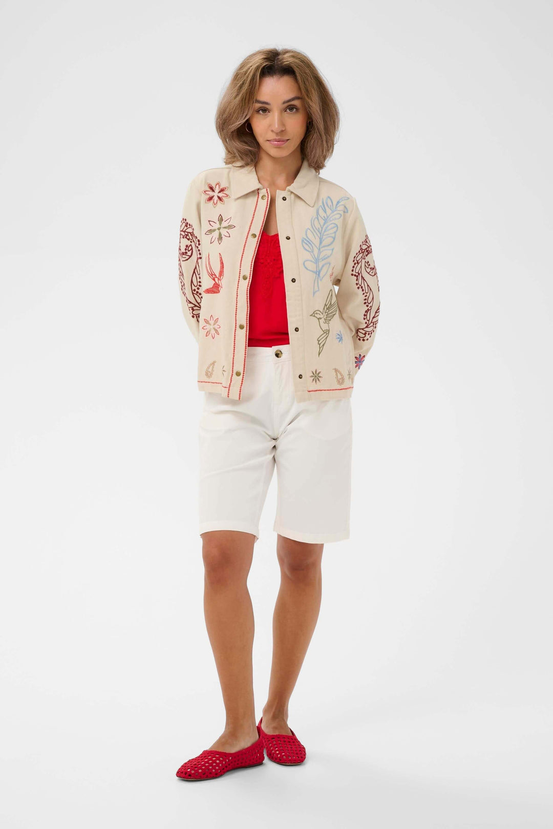 Femme portant une Veste Rosetta brodée à col chemise avec chemise rouge et shorts blancs, style élégant et confortable.