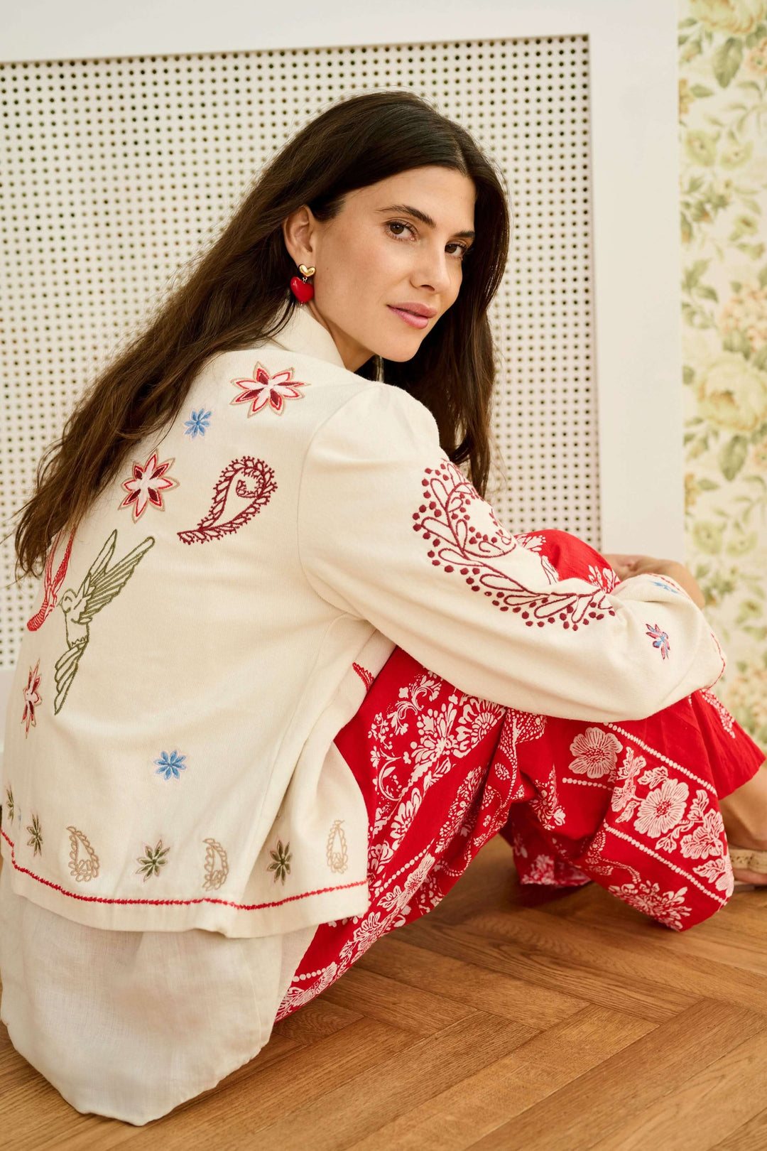 Femme portant une Veste Rosetta brodée à col chemise, avec des broderies artistiques colorées, assise sur un sol en bois, élégance décontractée.