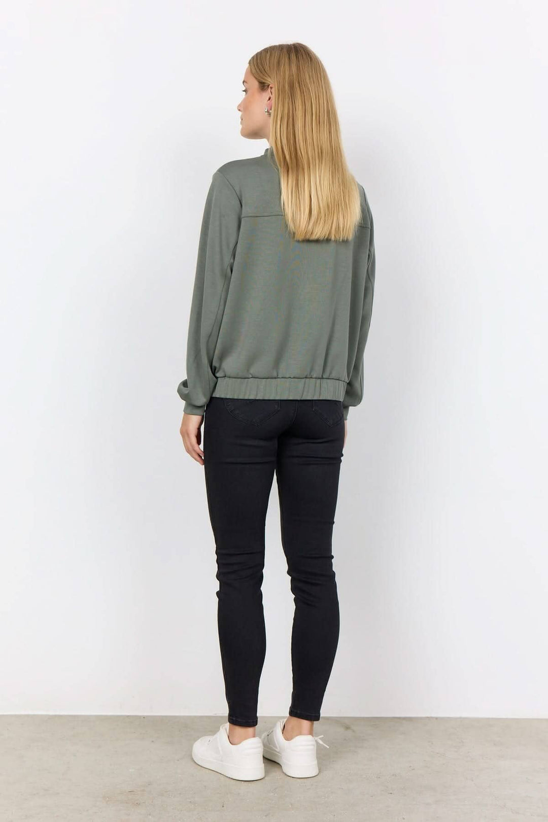 Femme portant une veste à bouton-pression verte, vue arrière, avec des pantalons noirs et des baskets blanches.