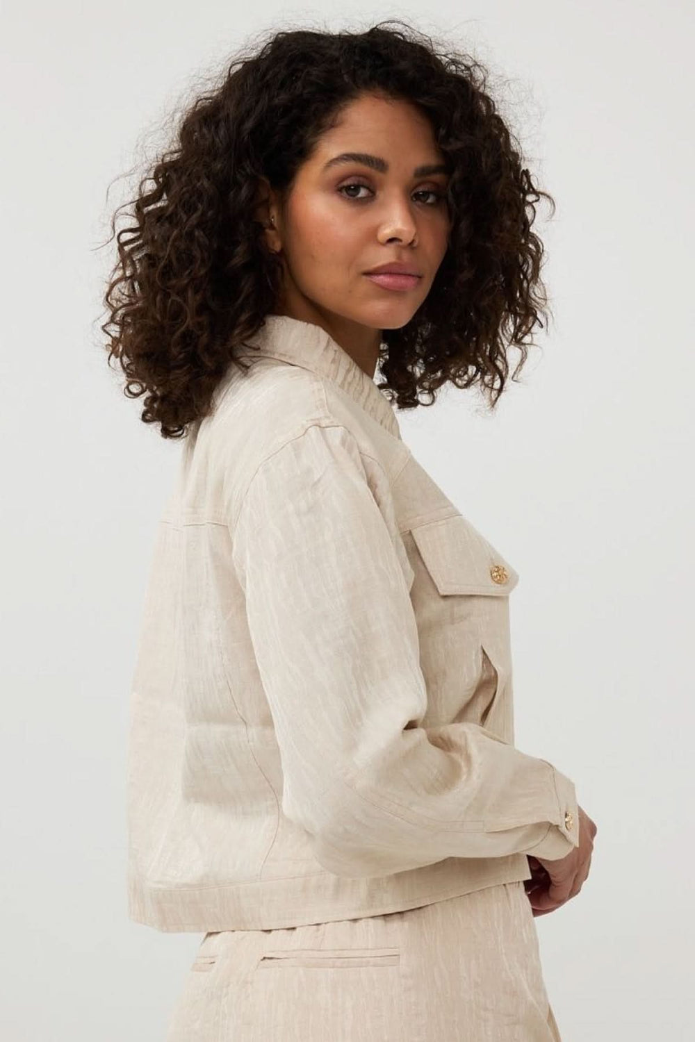 Veste courte en jacquard texturé beige avec poches à rabat et boutons dorés, portée par une femme aux cheveux bouclés.