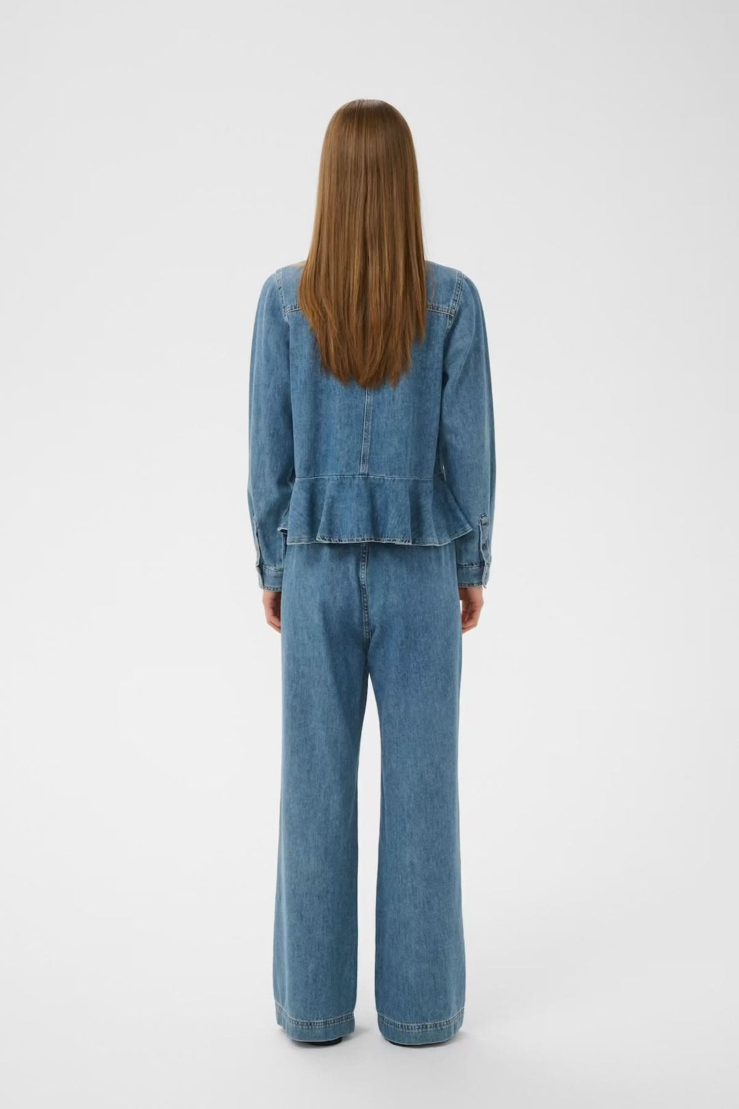Person wearing blue denim jacket and pants, standing with back to camera on white background.