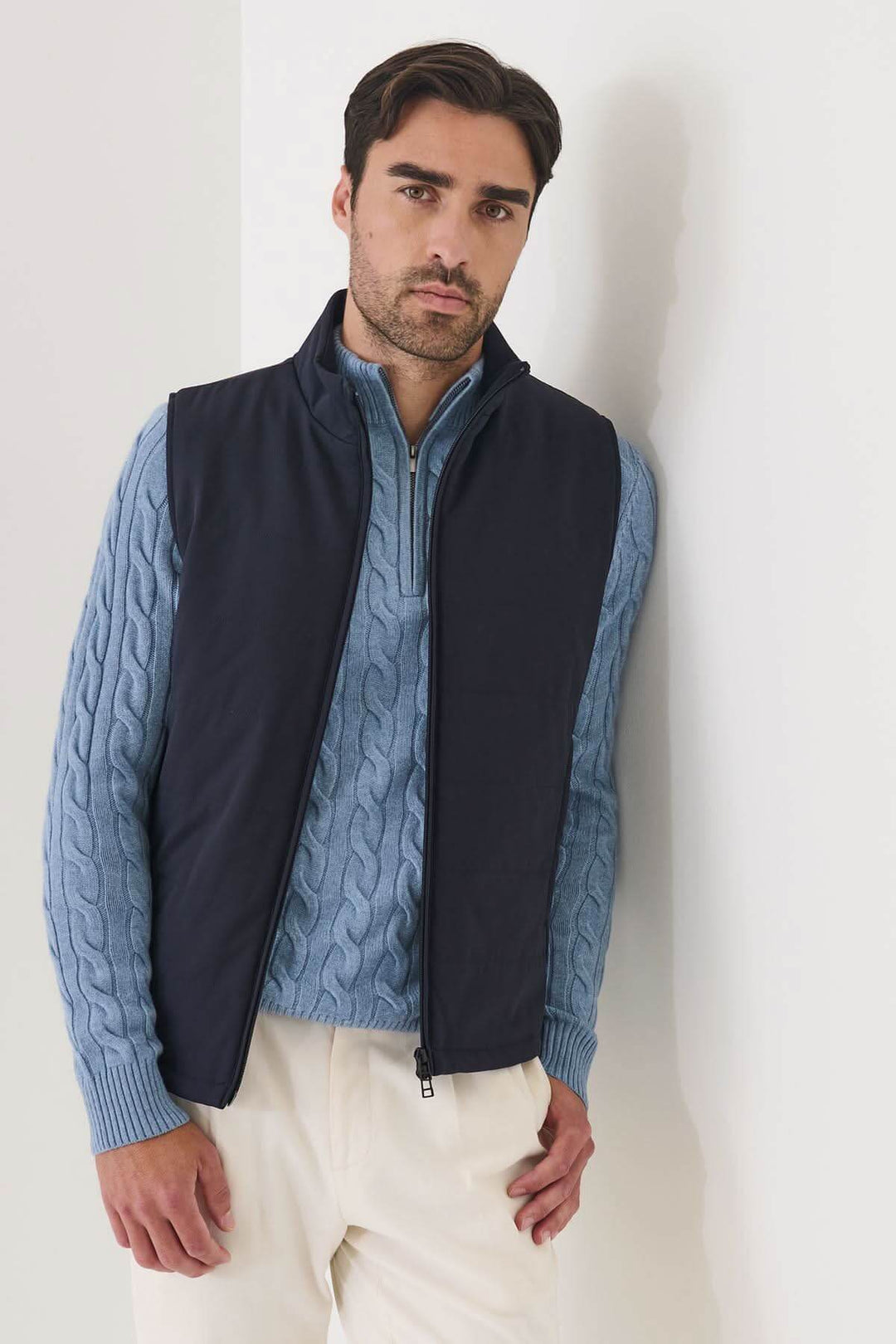 Homme portant la Veste sans manches matelassée Nucleo en tissu technique, avec pull bleu et pantalon blanc, posant contre un mur.