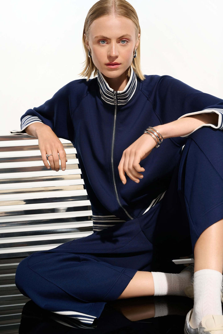 Femme portant une veste zippée à rayures contrastantes avec coupe droite et manches trois-quarts, assise sur un banc métallique.