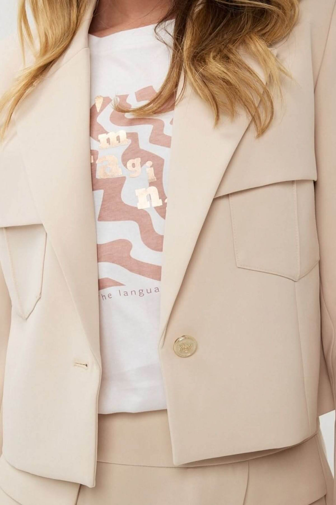 Veston court structuré en beige avec poches à rabat et bouton doré, porté sur un t-shirt graphique.
