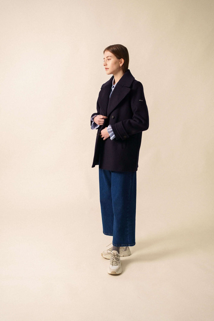 Femme portant un manteau chic noir avec col, encolure en V, et pantalon large, sur fond neutre.