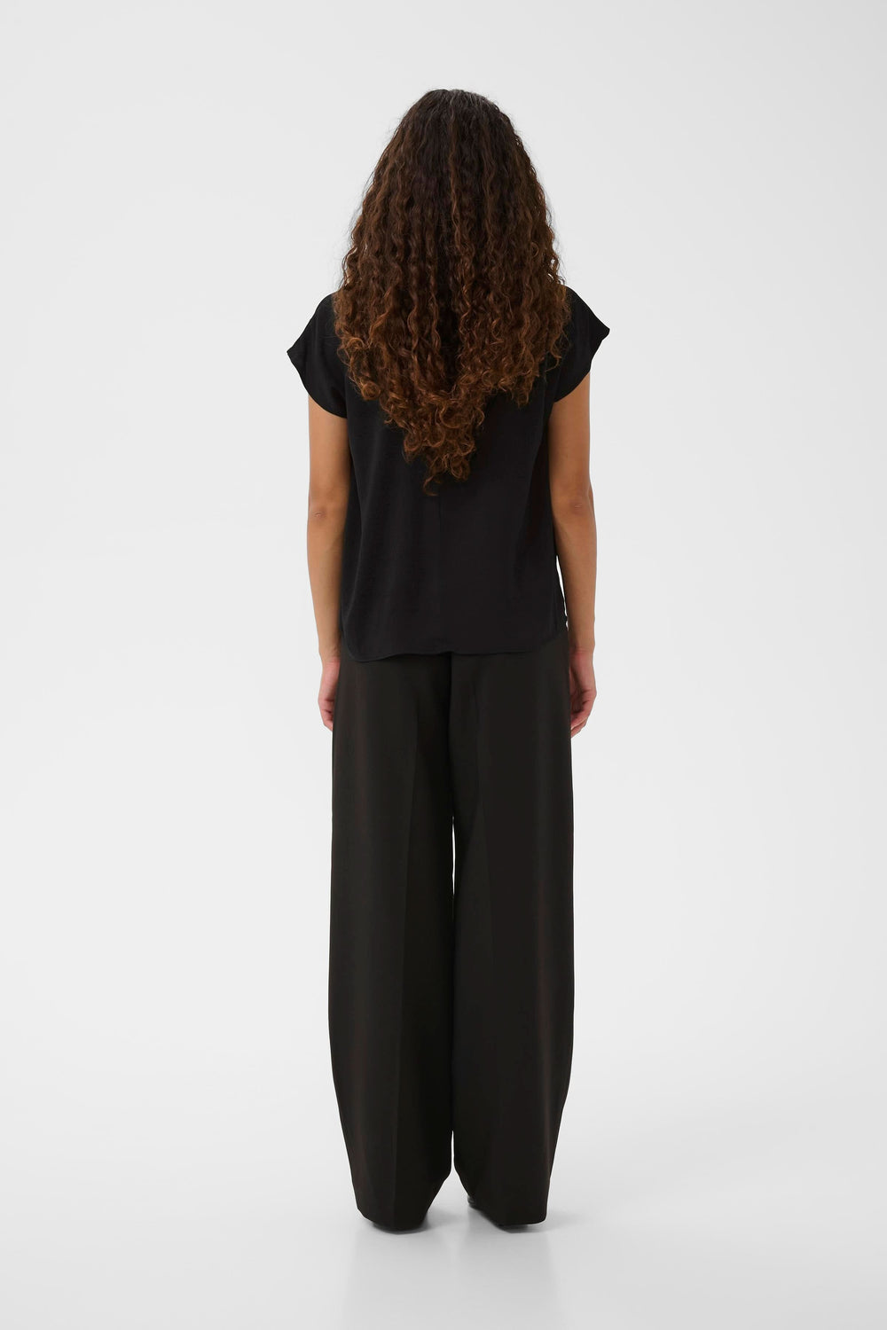 Vue arrière d'une femme portant un haut noir décontracté avec un pantalon ample, coiffée de cheveux bouclés.