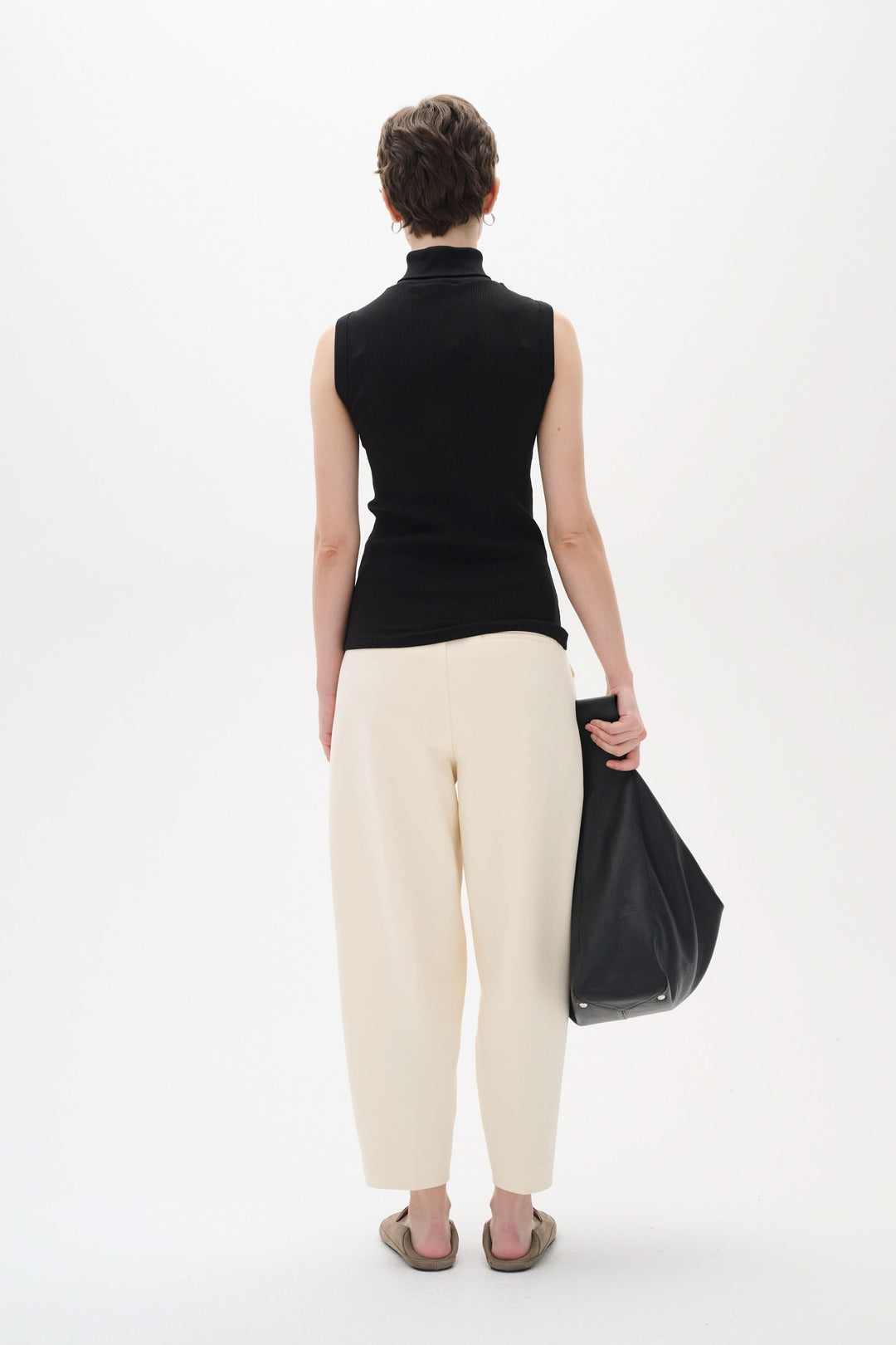 Femme portant une camisole noire sans manches et un pantalon crème, vue de dos avec un sac noir.