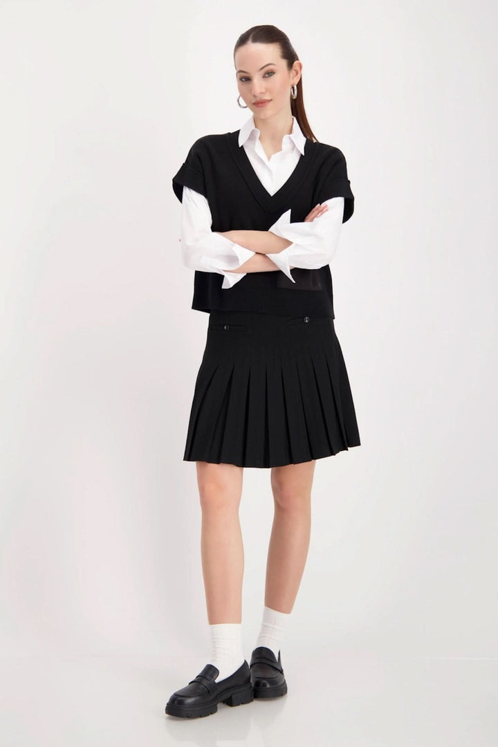 Femme portant un haut sans manches au col en V noir avec chemise blanche, style moderne et chic, jupe plissée, look tendance.