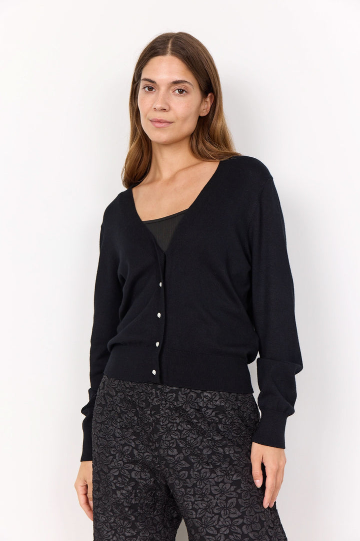 Cardigan classique noir à encolure en V, boutons brillants, porté avec un pantalon à motifs pour un look élégant.