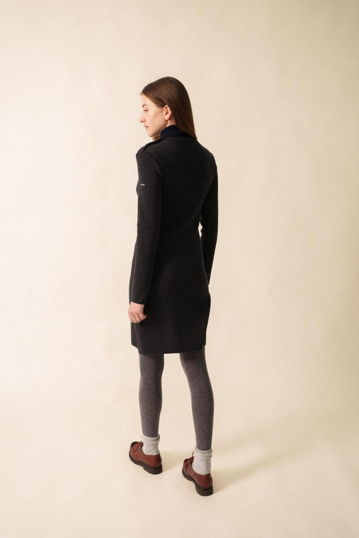 Femme de dos portant une robe ajustée en laine noire, avec des collants gris et des bottines marron.