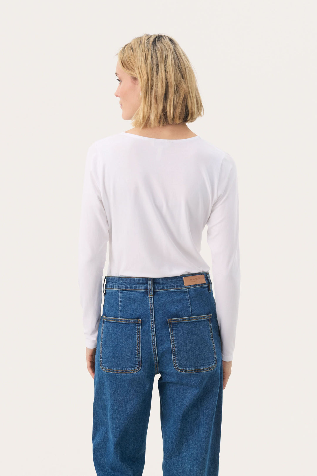 Femme vue de dos portant un chandail blanc à manches longues et un jean bleu, montré sur fond neutre.