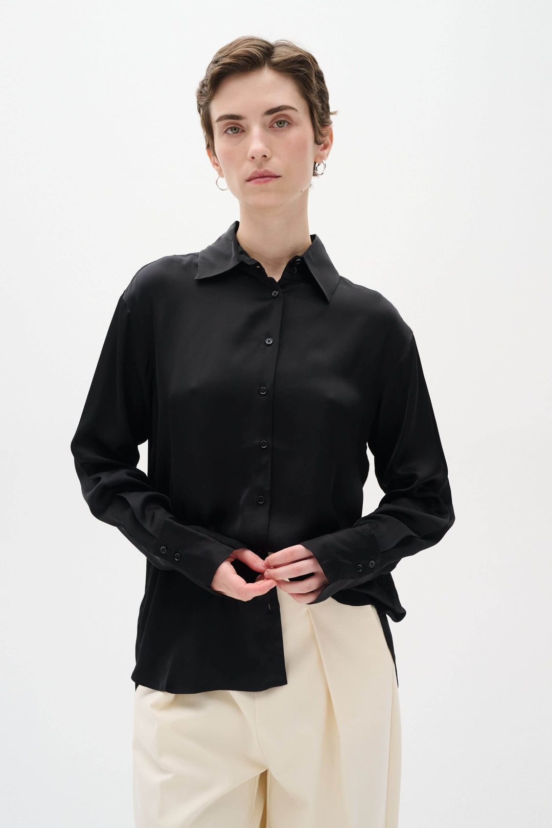 Chemise satinée noire Pauline à manches longues, coupe ample, portée avec un pantalon beige, style chic et décontracté.