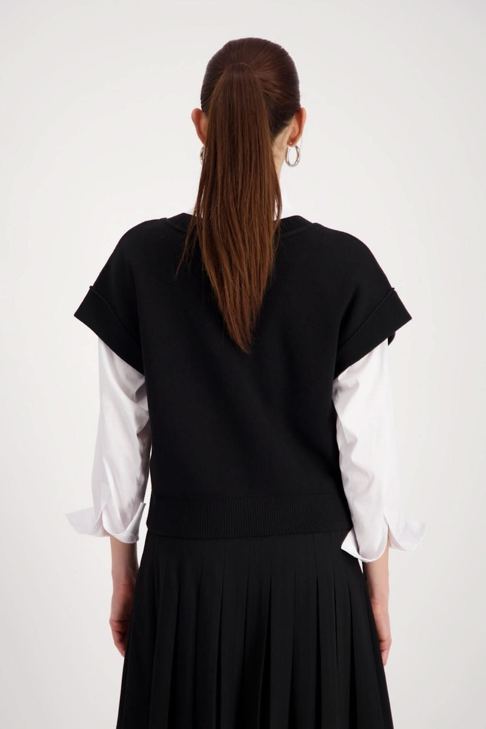 Femme portant un haut sans manches au col en V noir, superposé à une chemise blanche, vue de dos, avec jupe plissée.