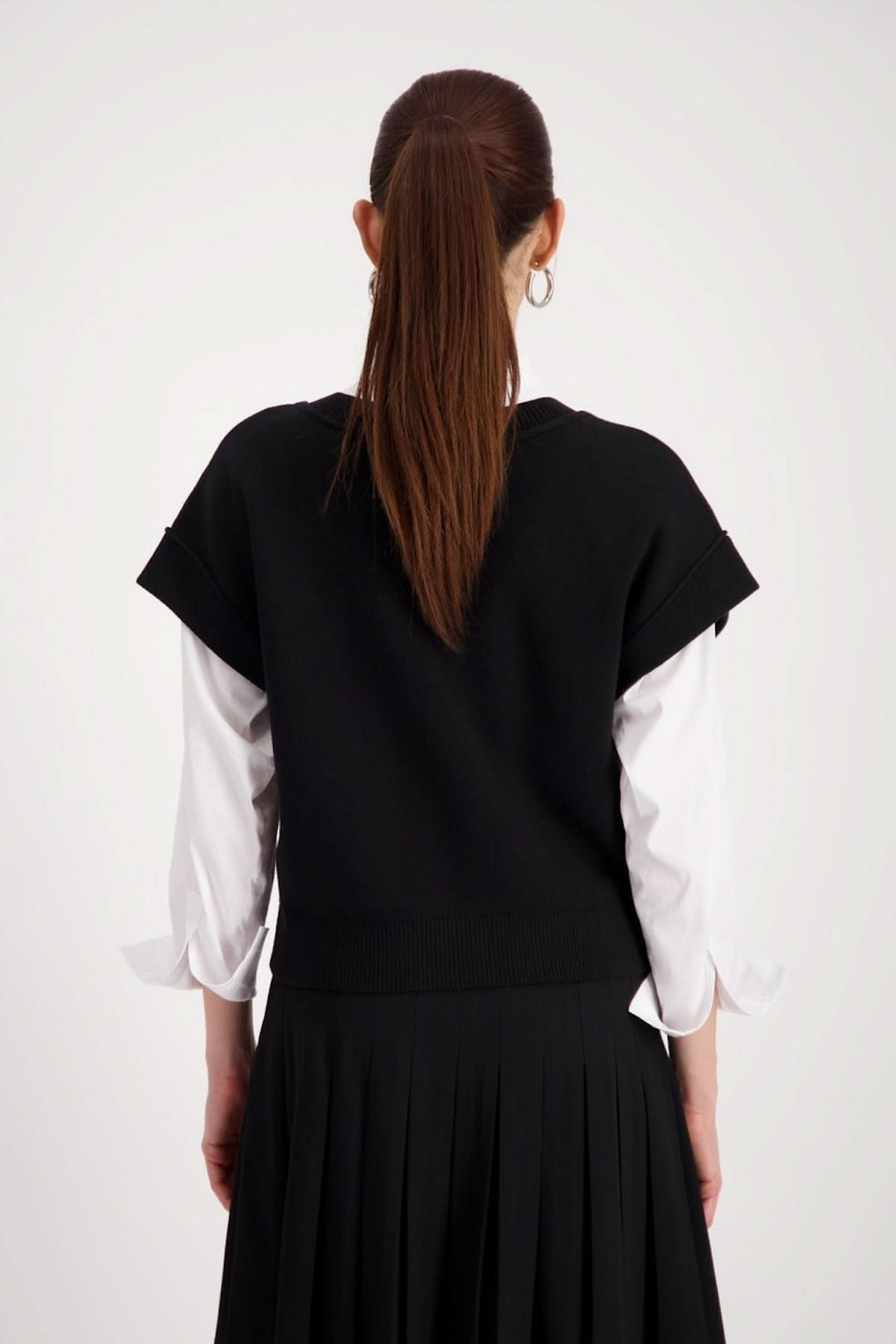 Femme portant un haut sans manches au col en V noir, superposé à une chemise blanche, vue de dos, avec jupe plissée.