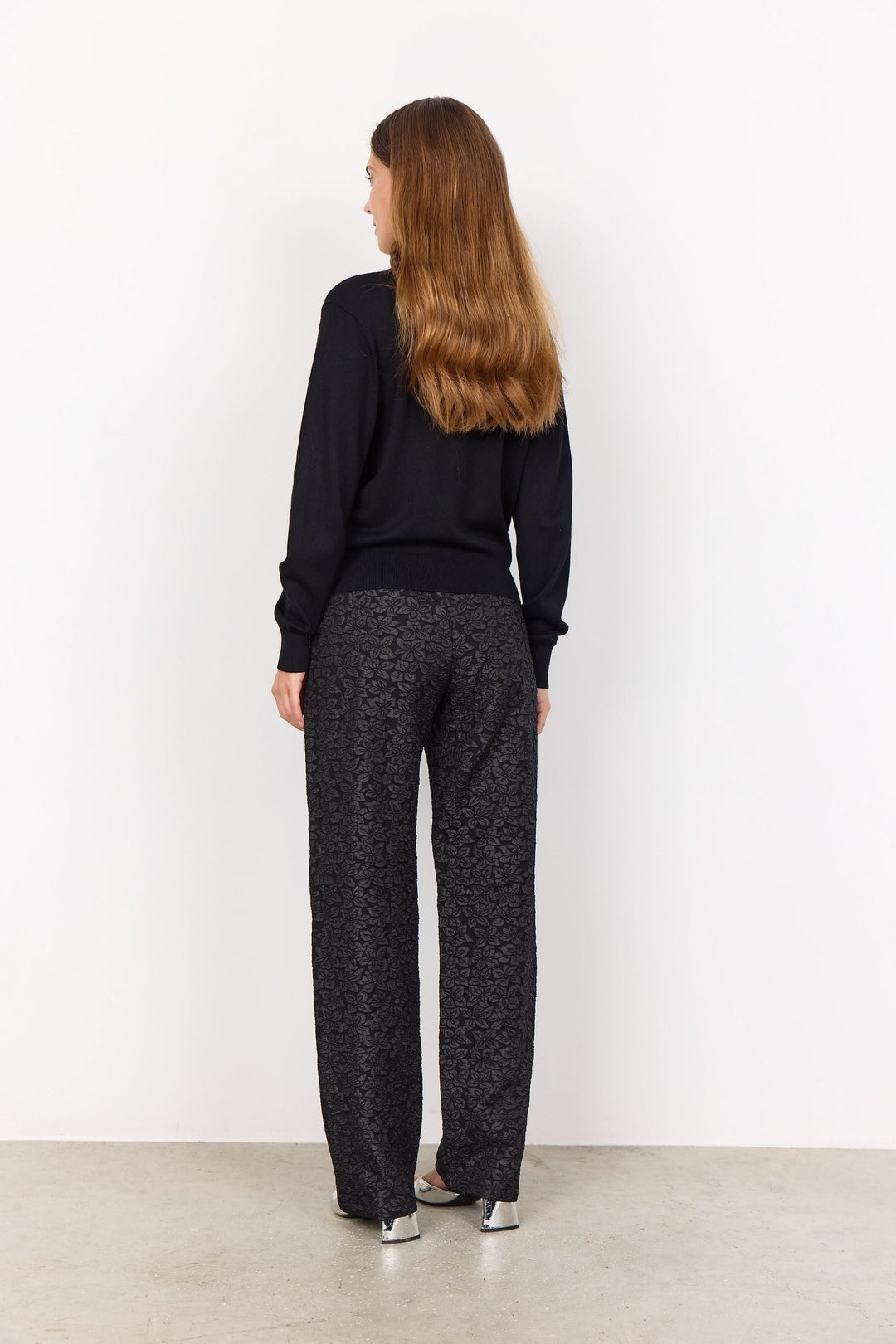 Femme portant un cardigan noir à encolure en V, vue arrière, avec un pantalon à motifs subtils.