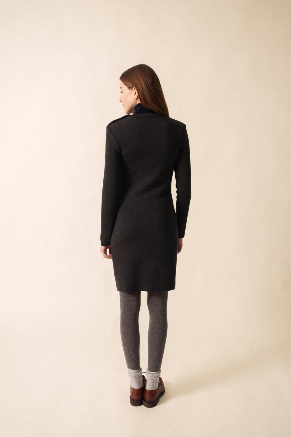 Femme de dos portant une robe ajustée en maille fine, avec collants et bottines, sur fond neutre.